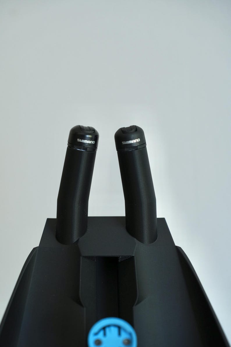 May include: Black bicycle handlebar grips with the brand name "Shimano" printed on the top. The grips are mounted on a black base. The image is taken against a white background.