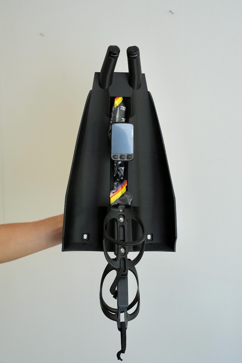 May include: Black bicycle accessory with two vertical handles at the top and a water bottle holder at the bottom. A small electronic device and a fuel packet are visible inside the accessory.