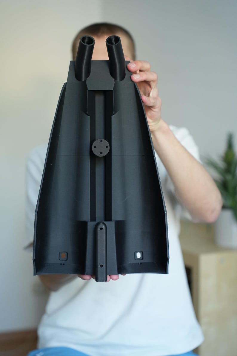 May include: A black, plastic component with two cylindrical nozzles and a central, elongated body. The component has a series of geometric cutouts and a circular attachment point. The object is held in front of a person.