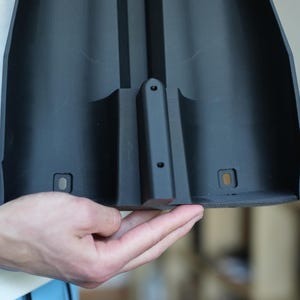 May include: A black plastic component with a curved design, featuring a central raised section and several small holes. The component has a matte finish and appears to be part of a larger structure.