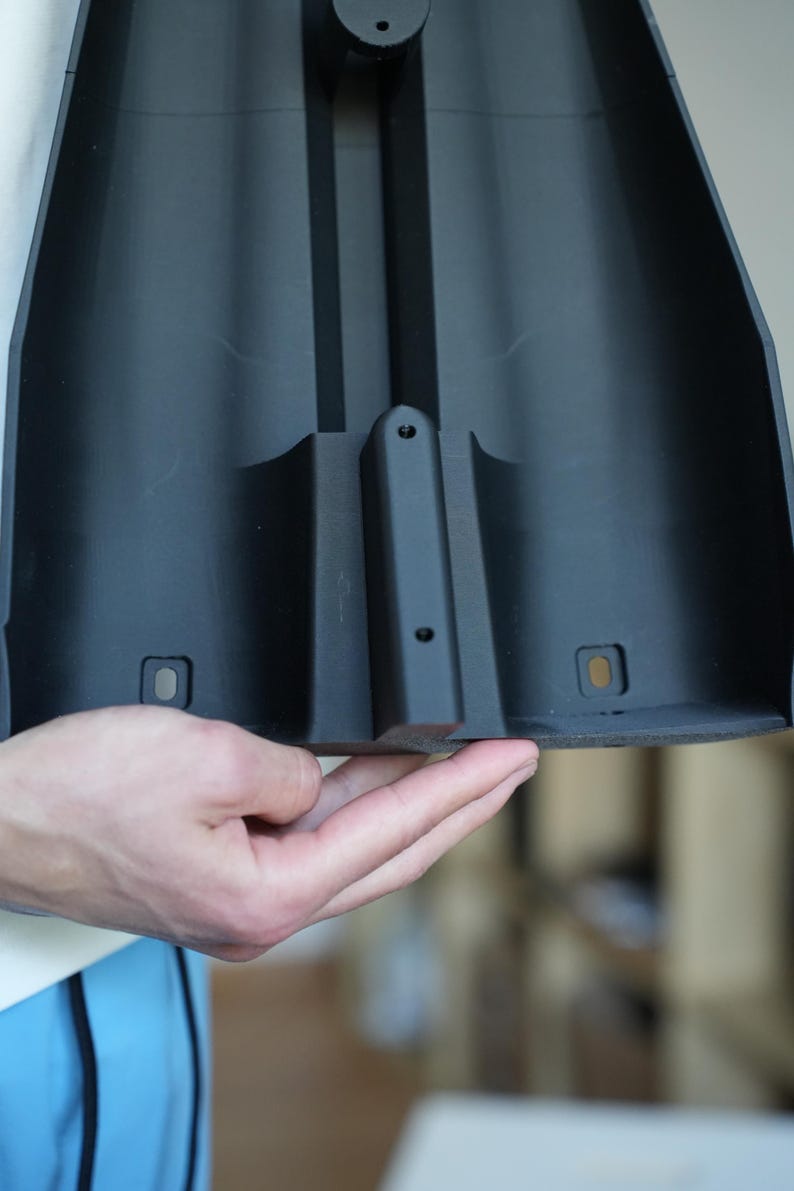 May include: A black plastic component with a curved design, featuring a central raised section and several small holes. The component has a matte finish and appears to be part of a larger structure.