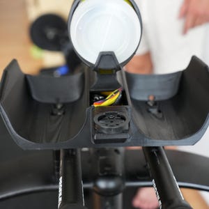 Pu&ograve; includere: Un supporto per prolunghe manubrio da bicicletta nero con due poggia-braccia curvi e un portaborraccia centrale. Il supporto include un attacco circolare nero per ciclocomputer e cavi visibili con accenti gialli e viola vicino al centro.