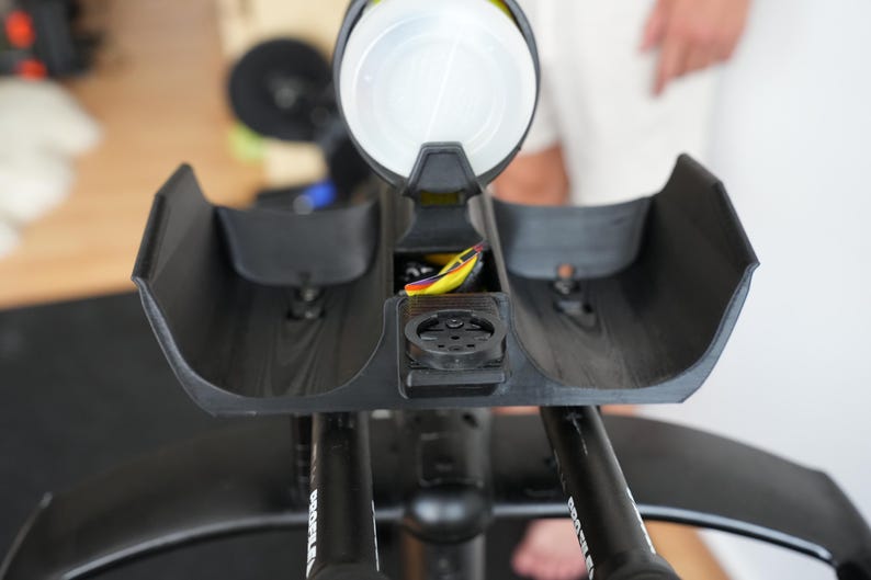 Pu&ograve; includere: Un supporto per prolunghe manubrio da bicicletta nero con due poggia-braccia curvi e un portaborraccia centrale. Il supporto include un attacco circolare nero per ciclocomputer e cavi visibili con accenti gialli e viola vicino al centro.
