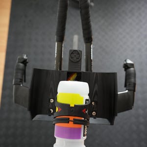 Pu&ograve; includere: Un set di appendici aerodinamiche nere per bicicletta con un portaborraccia centrale nero che sostiene una borraccia bianca con bande orizzontali gialle, arancioni e viola. Il montaggio include due barre di estensione nere con nastro adesivo.