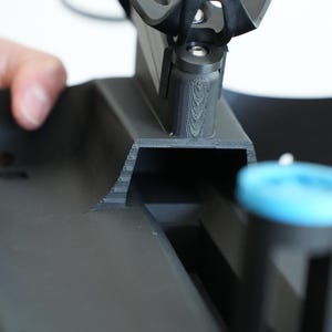 May include: Close-up of a black bicycle water bottle cage and mounting hardware. The cage is made of a curved, sturdy material designed to hold a water bottle. The image shows the cage's design and how it attaches to a bicycle frame.
