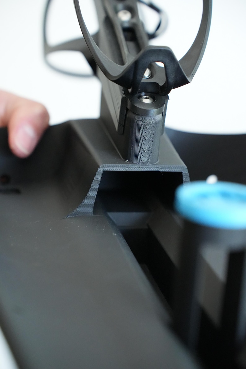 May include: Close-up of a black bicycle water bottle cage and mounting hardware. The cage is made of a curved, sturdy material designed to hold a water bottle. The image shows the cage's design and how it attaches to a bicycle frame.
