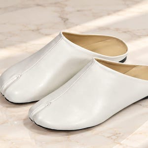 May include: A pair of white slip-on mules featuring a distinct split-toe design. These shoes have a smooth, solid white exterior, a beige interior lining, and a thin black sole. The front of each shoe is divided into two sections at the toe.