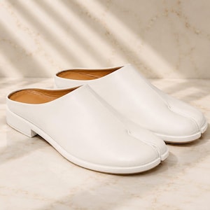 May include: A pair of white leather slip-on mules featuring a distinct split-toe design at the front. These shoes have a smooth finish, a low heel, and a tan-colored interior lining, showcasing a clean, minimalist construction with a rounded toe shape.
