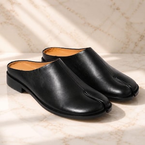 May include: A pair of black leather mules featuring a distinctive split-toe design, a low block heel, and a smooth finish. The slip-on footwear has a tan interior lining and a clean, rounded silhouette that covers the top of the foot.