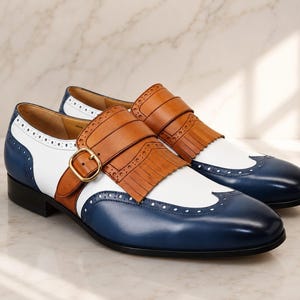 K&ouml;nnte beinhalten: Ein Paar Monk-Strap-Schuhe in dreifarbigem Design mit marineblauem Leder an Spitze und Ferse, wei&szlig;en Lederseitenteilen und einer beigen Lederfransenlasche &uuml;ber dem Spann. Die Schuhe verf&uuml;gen &uuml;ber eine goldfarbene Schnalle und Brogue-Verzierungen.