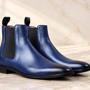 Könnte beinhalten: Ein Paar blaue Chelsea-Boots aus Leder mit glatter Oberfläche, dunklerem Farbverlauf an der abgerundeten Schuhspitze, schwarzen elastischen Seiteneinsätzen und schwarzen Zuglaschen an der Ferse. Die Stiefel haben einen flachen schwarzen Absatz.