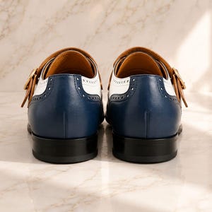 K&ouml;nnte beinhalten: Ein Paar Monk-Strap-Schuhe in dreifarbigem Design mit dunkelblauem Leder an der Ferse, wei&szlig;en Ledereins&auml;tzen und hellbraunen Lederriemen mit goldfarbenen Schnallen. Die Schuhe haben schwarze Abs&auml;tze und dekorative Brogue-Perforationen.