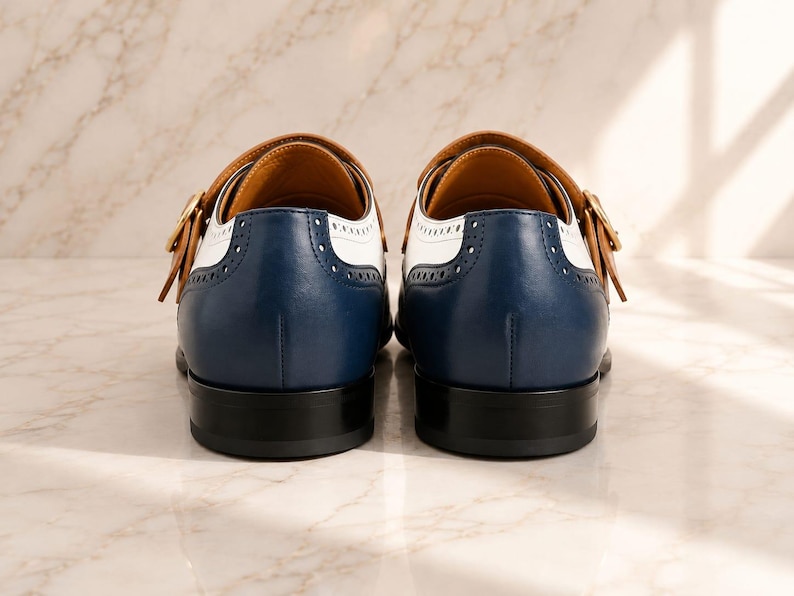 K&ouml;nnte beinhalten: Ein Paar Monk-Strap-Schuhe in dreifarbigem Design mit dunkelblauem Leder an der Ferse, wei&szlig;en Ledereins&auml;tzen und hellbraunen Lederriemen mit goldfarbenen Schnallen. Die Schuhe haben schwarze Abs&auml;tze und dekorative Brogue-Perforationen.