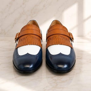 K&ouml;nnte beinhalten: Ein Paar Schnallenschuhe mit dreifarbigem Design, bestehend aus einer dunkelblauen Zehenkappe, einem wei&szlig;en Mittelteil und einer beigen Lederfransen-Applikation. Die Schuhe verf&uuml;gen &uuml;ber eine goldfarbene Schnalle am Riemen und Brogue-Lochmuster.