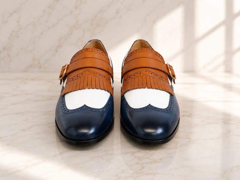 K&ouml;nnte beinhalten: Ein Paar Schnallenschuhe mit dreifarbigem Design, bestehend aus einer dunkelblauen Zehenkappe, einem wei&szlig;en Mittelteil und einer beigen Lederfransen-Applikation. Die Schuhe verf&uuml;gen &uuml;ber eine goldfarbene Schnalle am Riemen und Brogue-Lochmuster.
