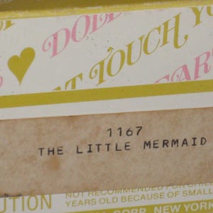May include: A close-up of a vintage box label featuring the text "THE LITTLE MERMAID" and the number "1167". The label is on a beige background with a yellow border and pink and yellow text.