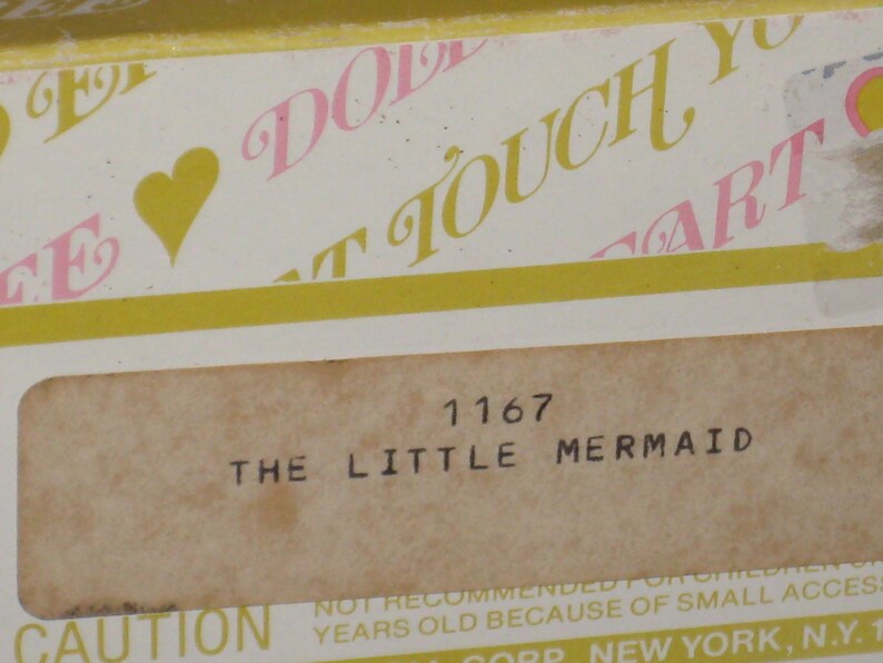 May include: A close-up of a vintage box label featuring the text "THE LITTLE MERMAID" and the number "1167". The label is on a beige background with a yellow border and pink and yellow text.