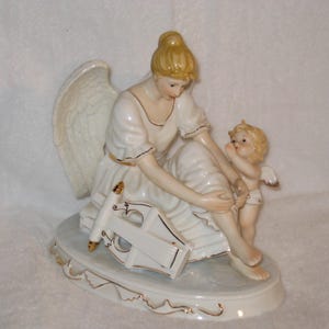 May include: A ceramic figurine of an angel with a cherub. The angel is seated, wearing a white dress with gold trim, and holding a lyre. The cherub stands beside her, with small wings. The base is white with gold accents.