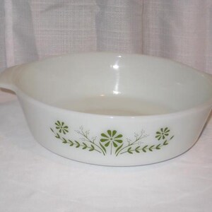 May include: A white milk glass round casserole dish with integrated side handles. The exterior features a green printed pattern of stylized flowers and leafy vines centered on the side of the bowl, creating a simple and clean aesthetic design.