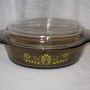 May include: An oval-shaped, smoke-colored glass casserole dish with a matching glass lid. The exterior of the dish features a bright yellow floral pattern consisting of daisies and leafy sprigs centered on the front side of the vessel.