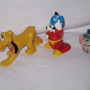 Puede incluir: Tres figuras de cer&aacute;mica de Disney: Pluto, Mickey Mouse y Dumbo. Pluto es amarillo con un collar negro y rojo. Mickey Mouse viste un atuendo rojo con un sombrero azul. Dumbo es gris con un sombrero azul y orejas rosas.