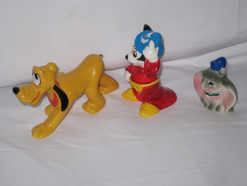 Puede incluir: Tres figuras de cer&aacute;mica de Disney: Pluto, Mickey Mouse y Dumbo. Pluto es amarillo con un collar negro y rojo. Mickey Mouse viste un atuendo rojo con un sombrero azul. Dumbo es gris con un sombrero azul y orejas rosas.