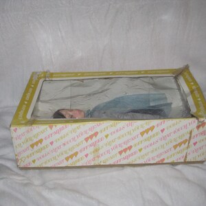 May include: A vintage doll in a box. The box is yellow with a clear plastic window and a printed pattern of hearts and text. The doll is visible through the window, lying down, wearing a blue dress.