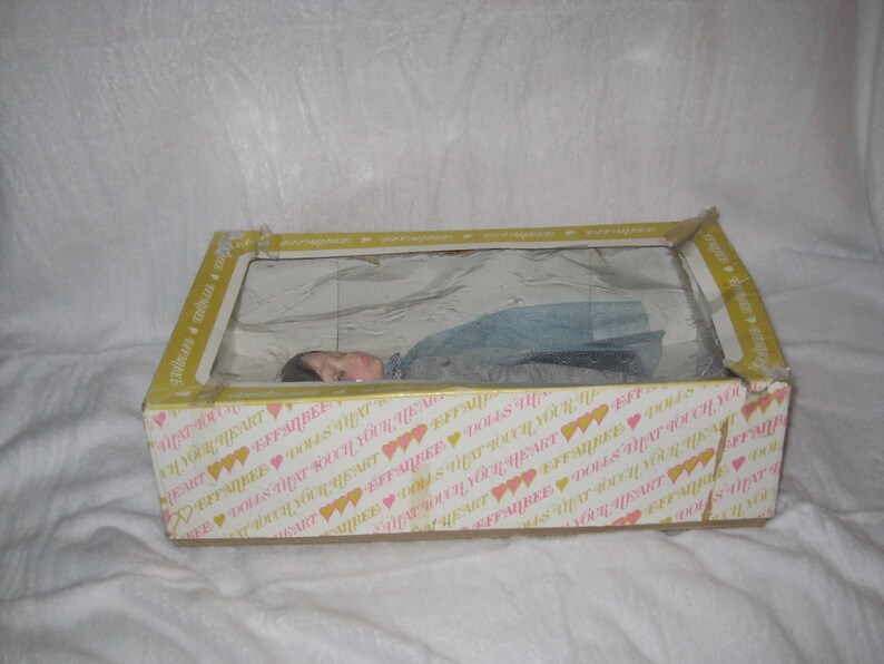 May include: A vintage doll in a box. The box is yellow with a clear plastic window and a printed pattern of hearts and text. The doll is visible through the window, lying down, wearing a blue dress.