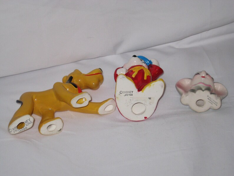 Puede incluir: Tres figuras de cer&aacute;mica: un perro amarillo, una figura en rojo y blanco y una figura de cara rosa. El perro amarillo est&aacute; tumbado. La figura roja y blanca tiene un sombrero azul. La figura rosa tiene orejas grandes.