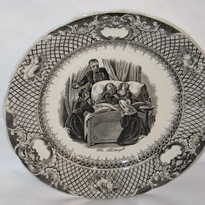 May include: A decorative plate with a black and white illustration depicting a scene with figures around a bed. The plate has a detailed border with floral and geometric patterns. The central image includes text.