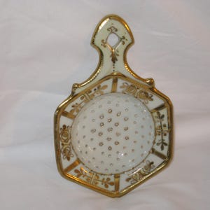 May include: An antique white porcelain trivet with gold trim and accents. The trivet has a handle and a central circular area with small, evenly spaced holes. The edges and handle are decorated with gold floral designs.