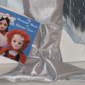May include: A silver doll with a mermaid-like tail, with a tag that reads "The Wonderful World of Effanbee Dolls." The tag features images of dolls. The doll's tail is made of a shiny, textured fabric.