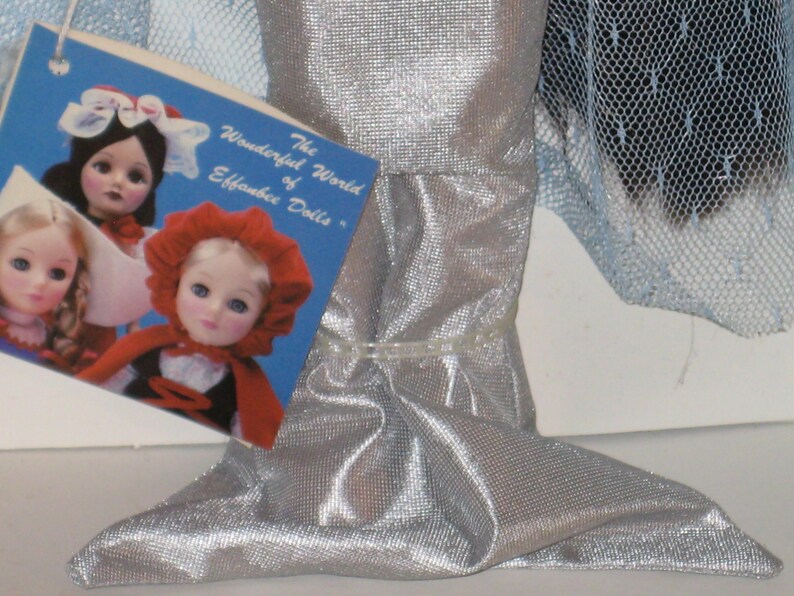 May include: A silver doll with a mermaid-like tail, with a tag that reads "The Wonderful World of Effanbee Dolls." The tag features images of dolls. The doll's tail is made of a shiny, textured fabric.