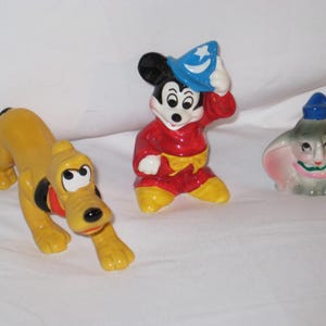 Puede incluir: Tres figuras de cer&aacute;mica: Pluto el perro en amarillo, Mickey Mouse con una t&uacute;nica roja y sombrero azul, y Dumbo el elefante con orejas rosas y sombrero azul. Las figuras est&aacute;n sobre una superficie blanca.