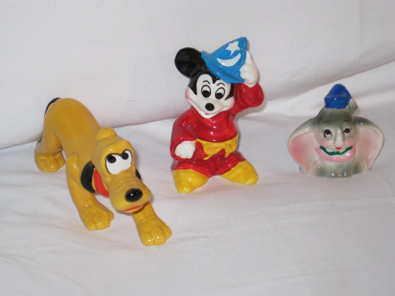 Puede incluir: Tres figuras de cer&aacute;mica: Pluto el perro en amarillo, Mickey Mouse con una t&uacute;nica roja y sombrero azul, y Dumbo el elefante con orejas rosas y sombrero azul. Las figuras est&aacute;n sobre una superficie blanca.