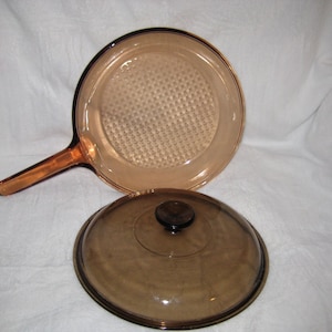 May include: A vintage amber-colored glass frying pan with a matching lid. The pan has a long handle and a textured cooking surface. The lid has a black knob. The cookware is designed for stovetop use.