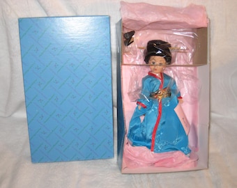 May include: A vintage doll in a box. The doll is dressed in a blue kimono with red trim and gold accents. The box is open, revealing the doll inside, and a separate blue box with a geometric pattern is next to it.