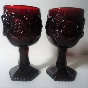 May include: Two ruby red goblets with ornate floral and geometric relief patterns. The stemware features a faceted base and a detailed design around the bowl. The glasses are made of a dark, translucent glass.