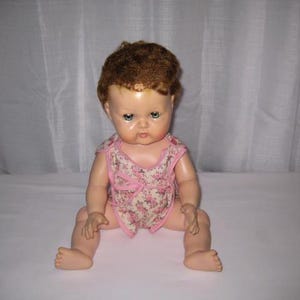 May include: A doll with short, curly reddish-brown hair and blue eyes. The doll has a light skin tone and is wearing a sleeveless, light-colored romper featuring a small pink floral pattern with pink trim around the neckline and arm openings.