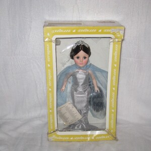 May include: A doll in a yellow box. The doll has dark hair, a silver dress, and a blue cape. The doll is holding a black purse. The box has the word "serinaires" printed on it.