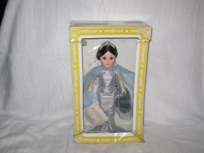 May include: A doll in a yellow box. The doll has dark hair, a silver dress, and a blue cape. The doll is holding a black purse. The box has the word "serinaires" printed on it.