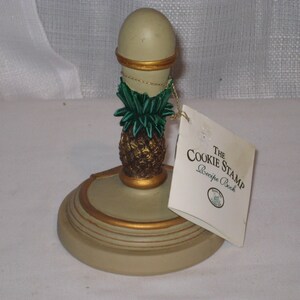 May include: A beige cookie stamp with a gold-painted circular base and a handle featuring a green and bronze pineapple design. A white paper tag is attached, printed with the text The Cookie Stamp Recipe Book and a small circular logo.