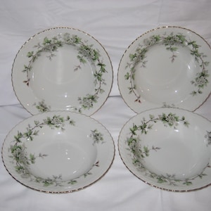 May include: Four white porcelain soup bowls with a floral design. Each bowl features a gold rim and a delicate pattern of green leaves and white flowers. The bowls are arranged on a white surface.