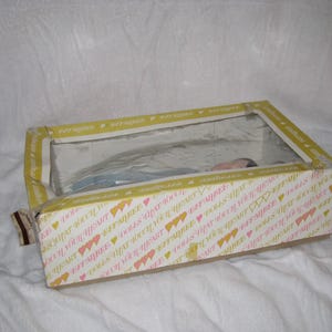 May include: A rectangular box with a clear plastic window, containing a doll. The box has a yellow and white pattern with hearts and text. The doll is partially visible through the window.