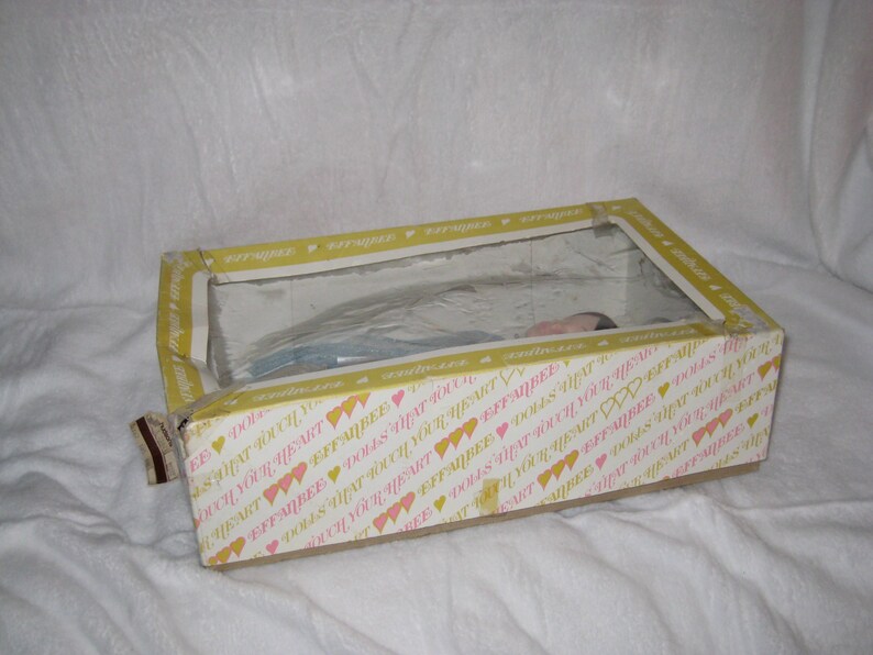 May include: A rectangular box with a clear plastic window, containing a doll. The box has a yellow and white pattern with hearts and text. The doll is partially visible through the window.