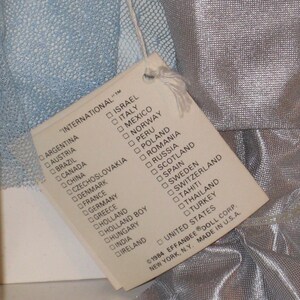 May include: A vintage doll tag listing various countries, including the UK, attached to a doll's silver fabric. The tag reads "INTERNATIONAL" and is from 1984. The tag is attached to a doll's silver fabric.