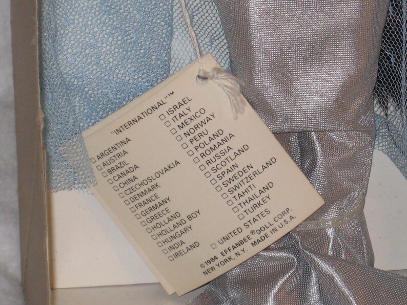May include: A vintage doll tag listing various countries, including the UK, attached to a doll's silver fabric. The tag reads "INTERNATIONAL" and is from 1984. The tag is attached to a doll's silver fabric.