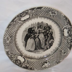May include: A decorative plate with a black and white illustration of a group of people in period clothing. The plate has a patterned border with floral and geometric designs. The central image depicts a scene with several figures, likely from a historical narrative.