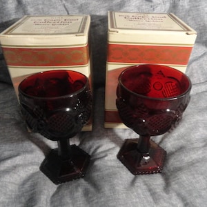 May include: Two deep red glass goblets from the Cape Cod Collection, each featuring a textured pattern on the bowl and a hexagonal base. The goblets are displayed in front of their original rectangular cardboard boxes with red and gold trim.