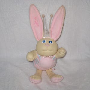 May include: A plush bunny doll with a cream-colored face and body, large pink ears, and blue eyes. The bunny wears a pink outfit with pastel stripes. The doll has a whimsical design, with a soft, cuddly appearance, and is likely a children's toy.